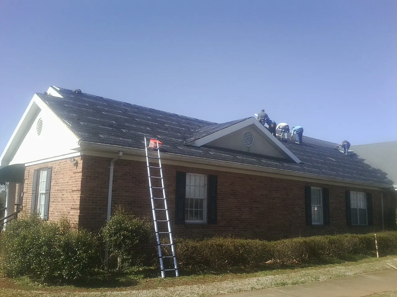 Crew tearing shingles off a brick ranch roof with a ladder set — Emergency Roof Repair work by our crew in Rhodesdale
