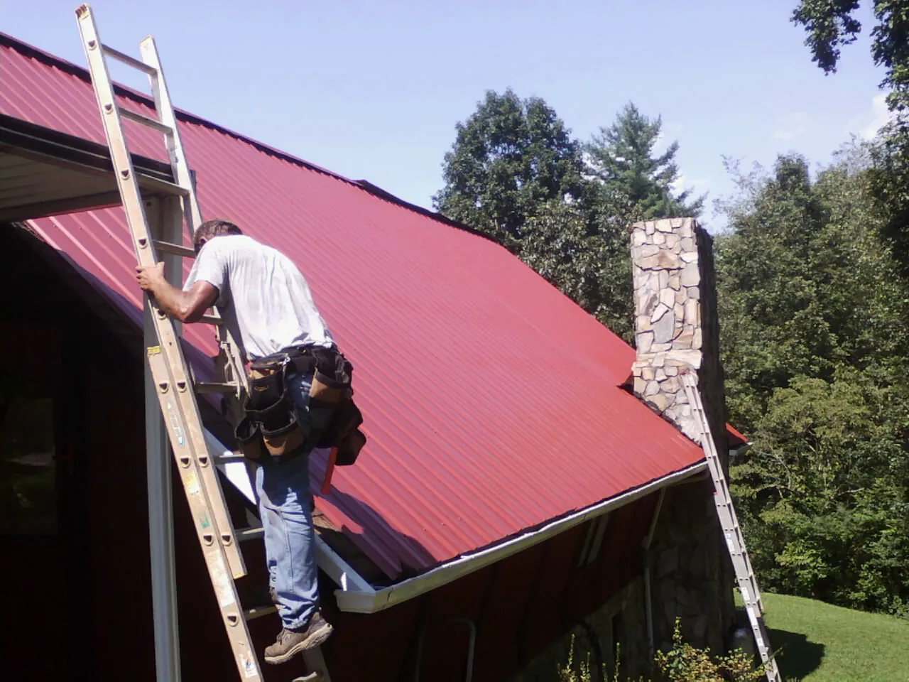 Roofer on a ladder beside a red metal roof and stone chimney — Soffit & Fascia Repair by Roofing Brothers in Rhodesdale